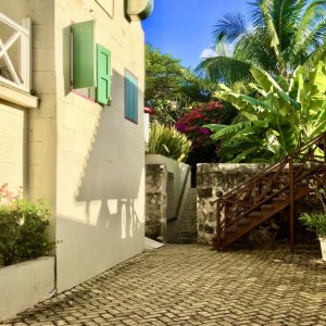 House with green shatters on a court with bougainvillae and palm trees and ground layers with brick stone